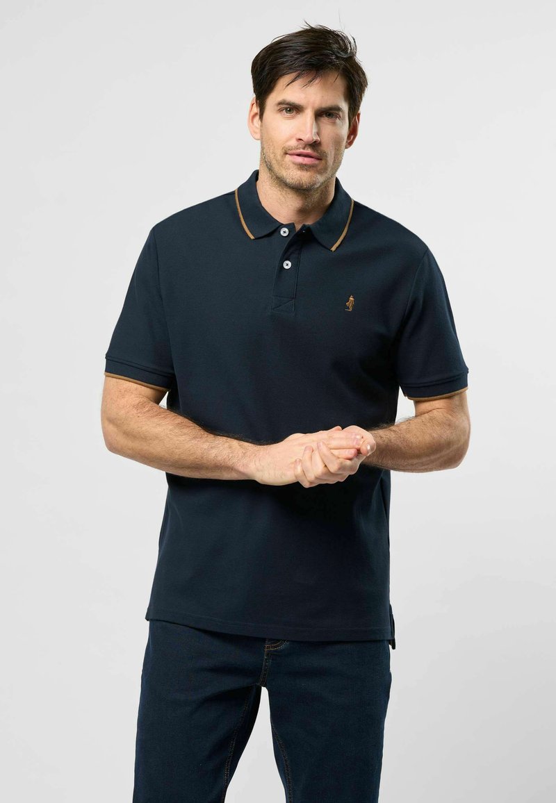 Homme aux cheveux courts et foncés, debout face à l’avant, portant un polo sombre avec bordure dorée et un jean bleu, sur un fond clair uni.