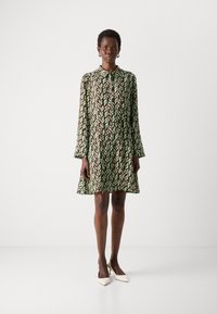 Abito a maniche lunghe di colore verde e marrone con motivo, colletto e orlo a campana. Indossato con décolleté bianchi a punta. Tessuto dalla texture liscia.