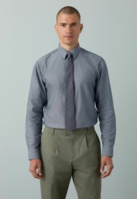 Graues Hemd mit langen Ärmeln und passender gemusterter Krawatte, kombiniert mit olivgrünen plissierten Hosen. Glatte Stofftextur.