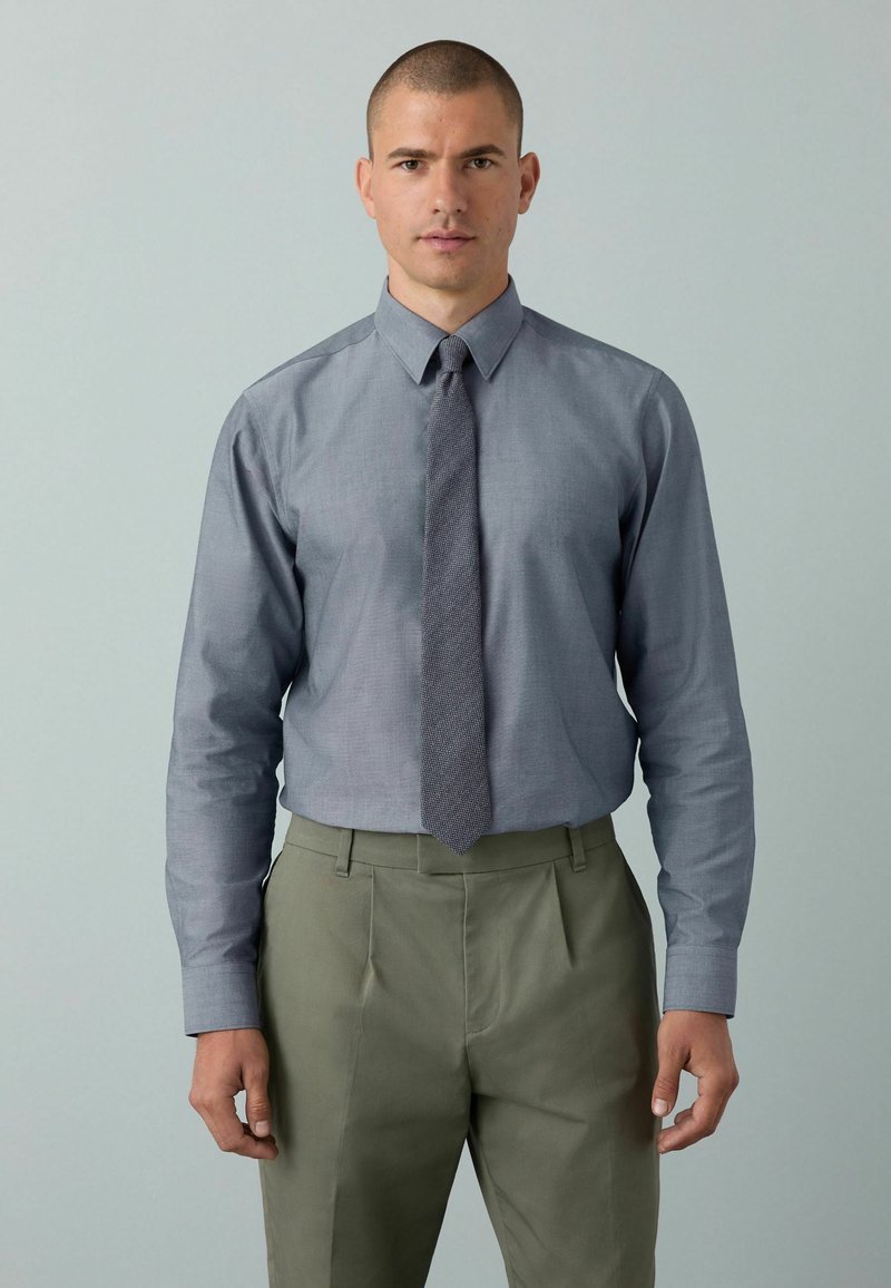 Graues Hemd mit langen Ärmeln und passender gemusterter Krawatte, kombiniert mit olivgrünen plissierten Hosen. Glatte Stofftextur.
