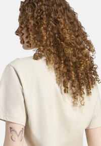 Camiseta de algodón color crema con mangas cortas, costuras visibles y un corte relajado, llevada por una persona con cabello rizado y un tatuaje de mariposa.