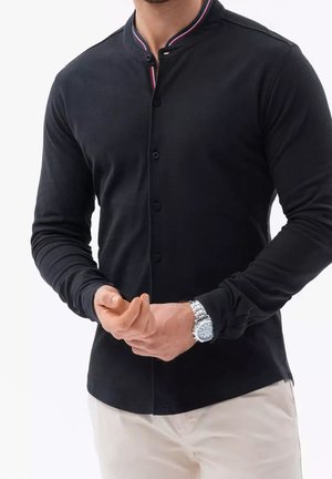 Mann trägt eng anliegendes schwarzes Hemd mit rot-weiß gestreiftem Kragen, beige Hose und silberne Armbanduhr, der den Ärmel anpasst.