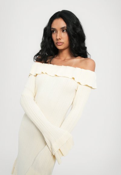 Off-the-shoulder ribbed sweater dress in soft cream with ruffled neckline and flared sleeve cuffs, featuring vertical texture throughout.
