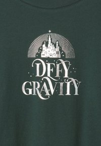 Sötétzöld anyag fehér grafikával, amely egy kastélyt, sugarakat és a "DEFY GRAVITY" szavakat díszes betűtípussal ábrázolja.