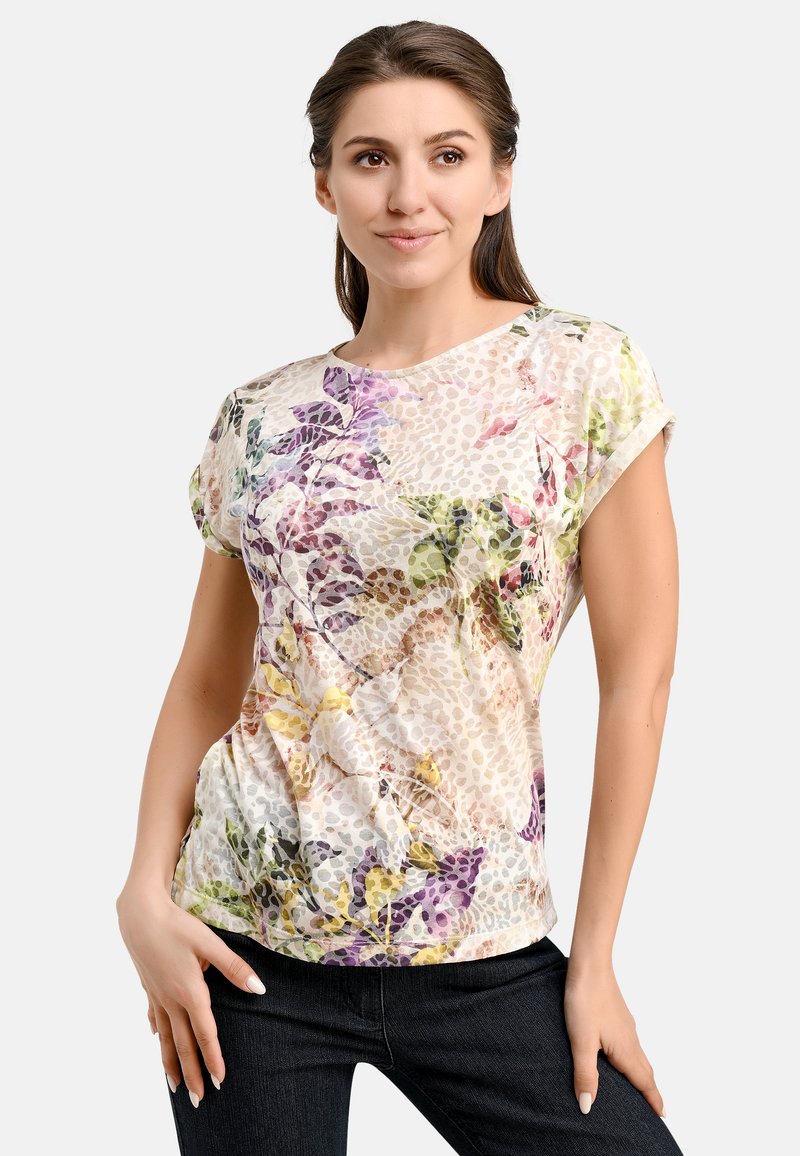 Bluse mit floralem Muster und kurzen Ärmeln aus hellcremigem Stoff. Sie hat lila und grüne Akzente, einen lässigen Schnitt, eine wellige Textur und einen runden Ausschnitt.