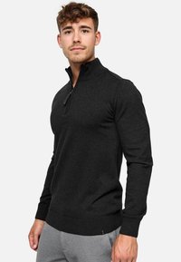 Junger Mann mit kurzen, lockigen Haaren, der einen eng anliegenden schwarzen Reißverschluss-Pullover und hellgraue Hosen trägt, vor einem schlichten weißen Hintergrund steht.