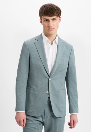 Junger Mann trägt einen hellblauen Zweiknopfanzug mit abgestepptem Revers und weißem Hemd, steht vor einem weißen Hintergrund.