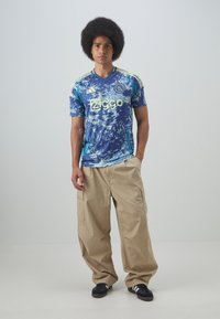 Maglia sportiva astratta blu e verde con logo giallo. Pantaloni cargo larghi color khaki, scarpe nere e un grande afro.