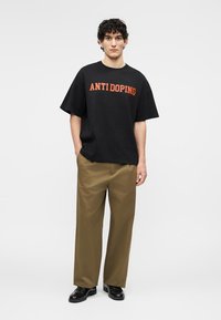 Jeune homme debout, les mains dans les poches, portant un t-shirt noir « ANTIDOPING », un pantalon marron à jambes larges et des chaussures noires, sur fond uni.