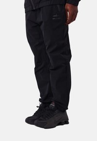 Pantalones impermeables negros con un ajuste holgado, bolsillos con cremallera y un sutil detalle de logo. Combinados con zapatos deportivos negros con detalles texturizados.