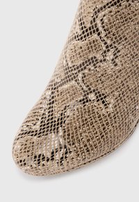 Chaussure beige à bout ouvert avec un design en maille et un motif dentelé, présentant un bout pointu et un revêtement intérieur noir. Surface texturée évidente.