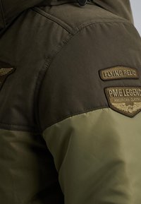 Grön och olivjacka med ett texturerat tyg. Har broderade lappar på axlarna, inklusive "Flying Relic" och "PME Legend."