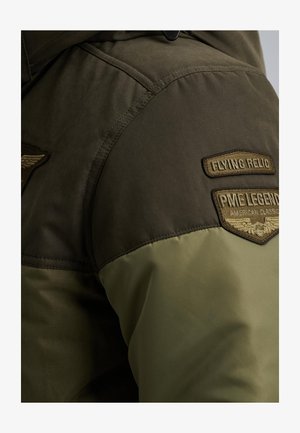 Grüne und olivfarbene Jacke aus strukturiertem Material. Verfügt über gestickte Patches auf der Schulter, darunter "Flying Relic" und "PME Legend."