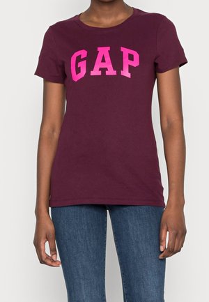 Person iført en kortærmet mørkerød T-shirt med lyserøde "GAP"-bogstaver og blå jeans, stående mod en ensfarvet baggrund.