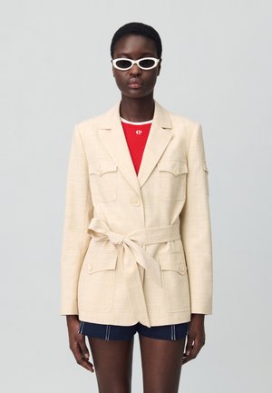 Blazer beige ceinturé avec deux poches avant, tissu texturé et col cranté. Associé avec un haut rouge et un short bleu marine. Lunettes de soleil en œil de chat blanches.