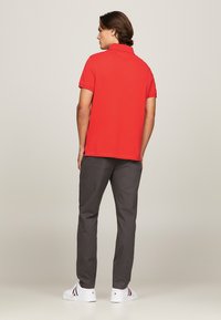 Röd kortärmad polotröja med klassisk krage, bärs med grå byxor och vita sneakers, sedd bakifrån. Slät tyg, avslappnad passform.