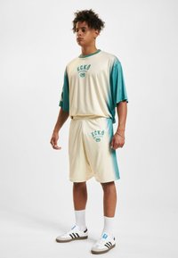 Outfit značky ECKO v krémovo-modrej gradientnej farbe; voľné tričko s krátkymi rukávmi, zodpovedajúce šortky a biele tenisky.
