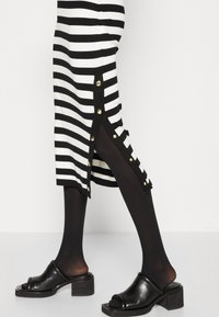 Jupe rayée noir et blanc longueur jambes avec des détails de boutons dorés sur la fente latérale, portée avec des collants noirs transparents et des chaussures à talon bloc ouvertes au bout.