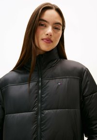 Schwarze Taillierte Jacke mit hohem Kragen, mit gesteppten Abschnitten und einem Reißverschluss. Kleines Logo-Detail auf der Brust. Glatte, glänzende Textur.