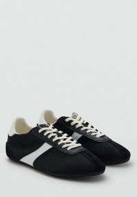 Par de zapatillas negras con rayas diagonales blancas, cordones blancos y detalles blancos en el talón sobre un fondo claro liso.