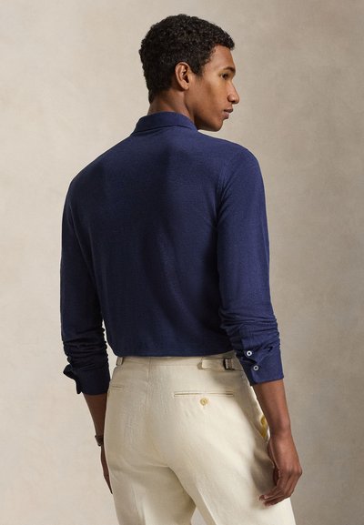 Άνδρας με κοντά σγουρά μαλλιά, που φορά εφαρμοστό μπλε σκούρο πουκάμισο με μακρύ μανίκι και εκρού ραμμένο παντελόνι, από πίσω.