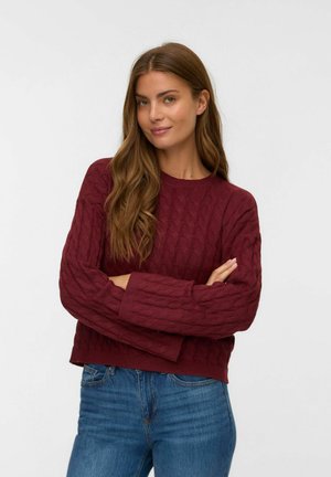 Femme aux longs cheveux bruns portant un pull en maille maroon et un jean bleu, les bras croisés, souriant légèrement sur un fond uni.