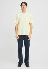 Kortermet, lysegul polo-skjorte med lomme. Flettet tekstur, flat krage og treknappelåpning. Kombinert med mørkeblå jeans.