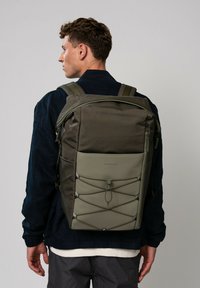 Sac à dos vert olive avec un tissu texturé, fermeture éclair, détail de cordon élastique à l'avant, bretelles réglables et un design épuré et structuré.