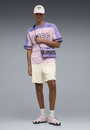Hombre con gorra blanca, camiseta deportiva en tonos morado y rosa con el número "0196", pantalones cortos color crema, bolso cruzado morado, calcetines blancos y zapatillas rosas de pie frente a un fondo gris.