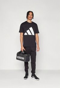 Juoda sportinė marškinėliai su dideliu baltu Adidas logotipu, derinami su juodomis kelnėmis ir juodais batais. Laiko juodą sportinę duffel tipo rankinę.