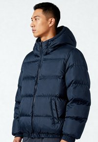 Giacca imbottita blu navy con cerniera frontale, cappuccio e polsini elasticizzati. Presenta sezioni trapuntate e una tasca laterale per riporre oggetti. Materiale resistente all'acqua.