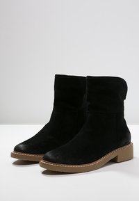 Bottines en suède noir avec un design légèrement affaissé, bout rond et semelle en caoutchouc beige. Présente des coutures visibles autour de la semelle.