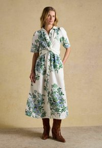 Femeie purtând o rochie midi albă cu imprimeu floral verde și albastru, mâneci scurte bufante, talie legată și cizme maro cu toc, stând în interior.