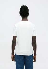 Mees, kellel on selja tagant vaadatuna lühikesed dreadlockid, kandes tavalist valget t-särki ja siniseid teksaseid valge tausta ees.