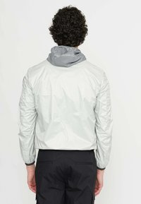 Letvægts, vandafvisende jakke i lys grå med en mørkegrå hætte. Har elastiske manchetter og en glat, shiny tekstur.