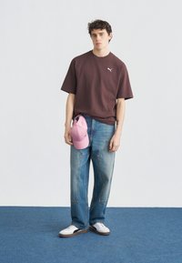 Νεαρός άνδρας που φοράει σκούρο καφέ t-shirt Puma, μπλε τζιν, λευκά αθλητικά παπούτσια, κρατάει ένα ροζ καπέλο Puma, στέκεται πάνω σε μπλε χαλί μπροστά σε ανοιχτόχρωμο τοίχο.