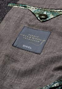 Brauner Stoff mit texturierter Oberfläche, der ein grün gemustertes Akzentdetail und ein Etikett mit der Aufschrift "Finest Silkwool Selection, Digel" trägt.