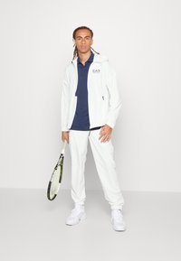 Balta sportinė striukė ir kelnės su tamsiai mėlyna polo marškinėliais, laikantis teniso raketę. Striukė turi logotipą, tekstūruotą audinį ir sportinius batus.