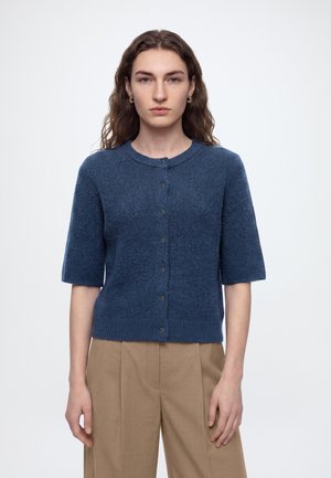 Kvinde med langt bølget hår iført en blå strikket sweater med korte ærmer og beige bukser med høj talje, stående mod en ensfarvet baggrund.