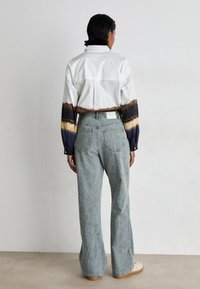 Persona con cabello corto y oscuro se encuentra de pie frente a una pared blanca, vistiendo una camisa blanca con bandas en las mangas de color marrón y azul, jeans azules con estampado y zapatos blancos.