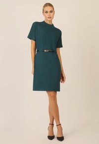 Vestido azul verdoso con mangas cortas, cuello alto y un cinturón negro con hebilla dorada; ajustado con abertura lateral y textura suave. Tacones negros.