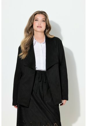 Frau mit langen, welligen Haaren trägt ein weißes Hemd mit Knöpfen, einen schwarzen offenen Blazer und einen schwarzen Rock mit floralem Cutout-Muster am Saum.