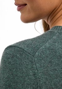Maglione verde lavorato a maglia con una texture morbida, cuciture visibili e un collo rotondo, mostrato da dietro, evidenziando la cucitura della spalla.