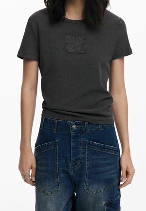 Femme portant un t-shirt à manches courtes gris foncé avec les lettres "DSGL" et un jean bleu foncé taille haute avec de grandes poches avant.