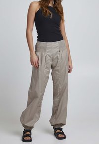 Light beige, loose-fitting trousers with a smocked waistband and gathered texture. Paired with a black tank top and black sandals.