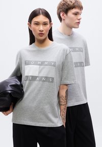 Γκρι βαμβακερά T-shirt με κουτί σε γραμμή, με λευκό και γκρι γράφημα "TOMMY JEANS" στο μπροστινό μέρος. Κοντά μανίκια, λεία υφή.