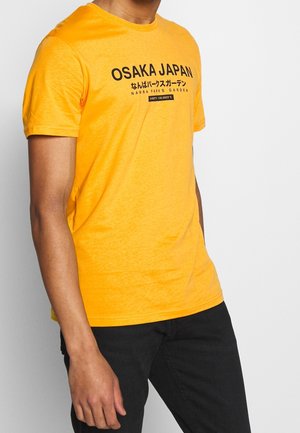 Muž v jasně oranžovém tričku s nápisem "Osaka Japan" a japonským textem na přední straně, v kombinaci s černými kalhotami.