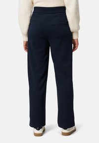 Marineblå bukser med vide ben og glat tekstur, der har to baglommer og en høj talje, kombineret med hvide sneakers.