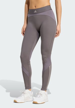 Donna che indossa leggings grigi Adidas e scarpe da ginnastica bianche, in piedi dritta contro uno sfondo chiaro e semplice.
