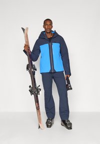 Sí öltözet, amely egy kék és sötétkék dzsekiből áll, cipzárakkal, sötétkék nadrágból, fekete sícipőkből és fekete hardverrel ellátott fából készült sílécekből.